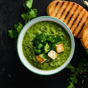 Kom met groene broccoli-soep, gegarneerd met croutons en verse kruiden, naast geroosterd brood op een donkere ondergrond